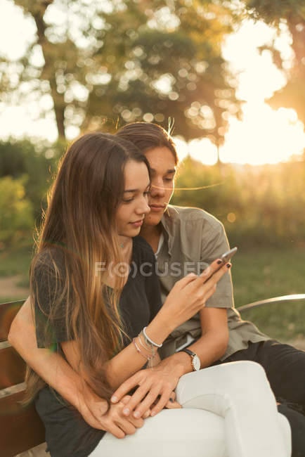 Ritratto di ragazzo che abbraccia la fidanzata navigando smartphone in panchina nel parco — Foto stock