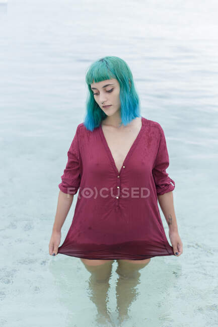 Portrait of sad young blue haired girl standing in water, looking down and holding wet red shirt. — Stock Photo
