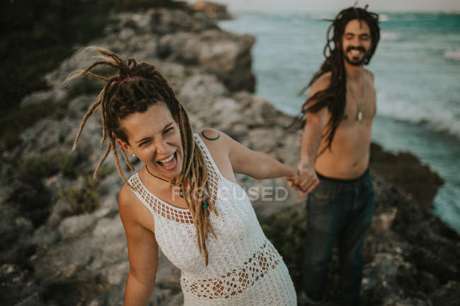 Retrato de la alegre chica y el hombre tomados de la mano y riendo en las rocas en la costa del océano - foto de stock