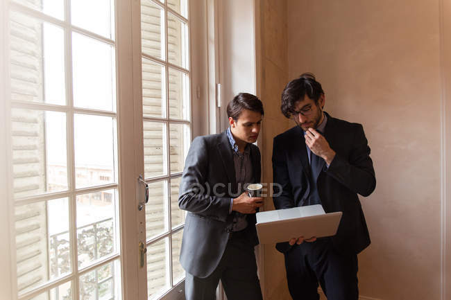 Zwei junge Geschäftsleute stehen am Fenster und schauen auf Laptop. — Stockfoto