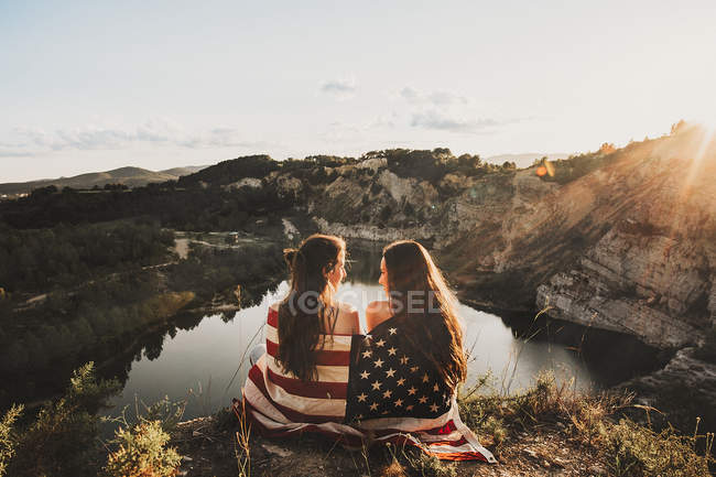 Вид сзади на двух девушек, завернутых в американский флаг, сидящих у отдаленного горного озера — стоковое фото