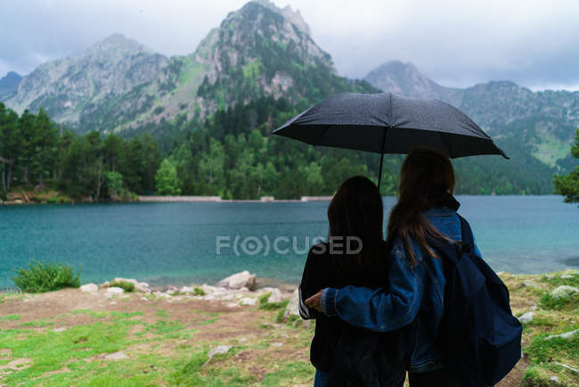 Niñas bajo paraguas de pie en la orilla del lago de montaña - foto de stock
