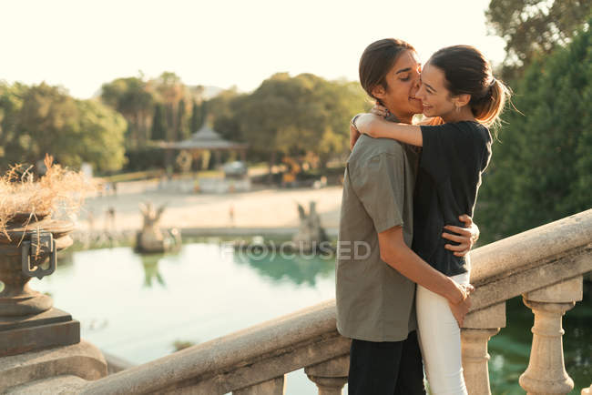 Retrato de namorado abraçando namorada e beijando na bochecha nas escadas no parque sobre lago no pano de fundo — Fotografia de Stock