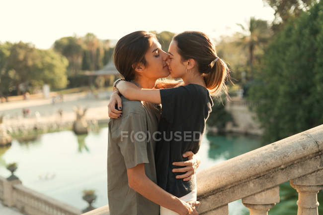 Retrato de namorado abraçando namorada e beijando no nariz nas escadas no parque sobre o lago no fundo — Fotografia de Stock