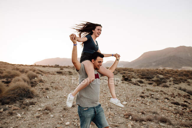 Щаслива молода пара сміється і посміхається в сільській місцевості проти гір і блакитного неба. Чоловік тримає жінку на плечах . — стокове фото