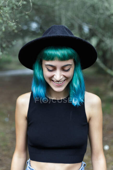 Закрыть вид привлекательной молодой девушки-хипстера с ярко-синими волосами, стоящей в парке, глядя вниз и улыбаясь. — стоковое фото