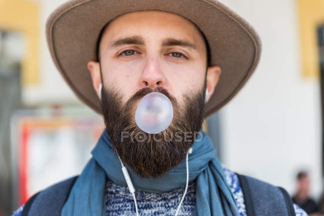 Barbuto uomo in cappello guardando la fotocamera e soffiando gomma bolla . — Foto stock