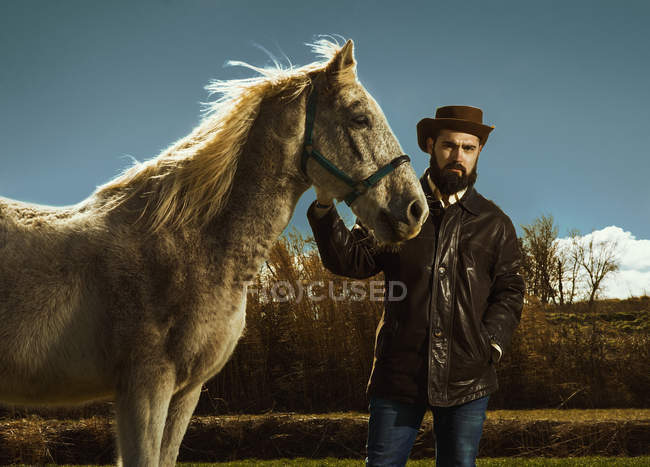 Homem barbudo segurando cavalo e olhando para a câmera no campo — Fotografia de Stock