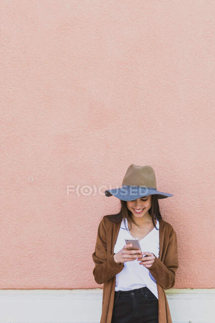 Porträt eines lächelnden Mädchens mit Hut, das sein Handy an der rosafarbenen Wand kontrolliert — Stockfoto