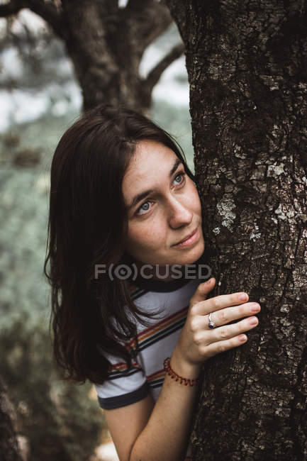 Брюнетка жінка фотографіях хтось дивитися вбік при, ховаючись за стовбур дерева — стокове фото