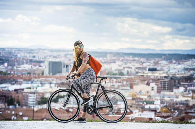 Side view portrait of a young woman with tattooed arm riding bike in city street. — Stock Photo