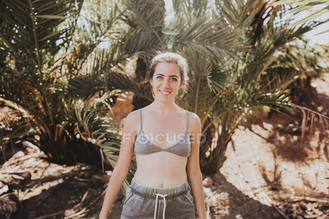 Fille gaie en haut de bikini souriant à la caméra contre des palmiers — Photo de stock