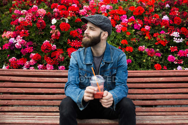 Portrait of bearded man sitting on bench with smoothie on background of rose bushes — Stock Photo