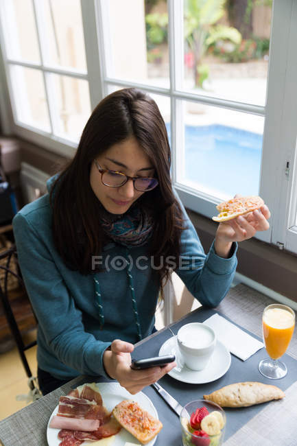 High angle portrait of woman in eyeglasses having toast and browsing smartphone at table in cafe — Stock Photo