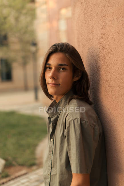 Retrato de menino com cabelos longos apoiados na parede marrom e olhando para a câmera — Fotografia de Stock