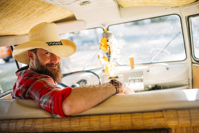 Man in cowboy hat sitting at drivers seat of retro van and looking at camera — Stock Photo