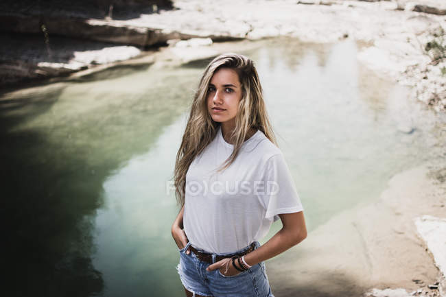 Молода жінка стоїть на озері з руками в кишенях і дивиться на камеру — стокове фото