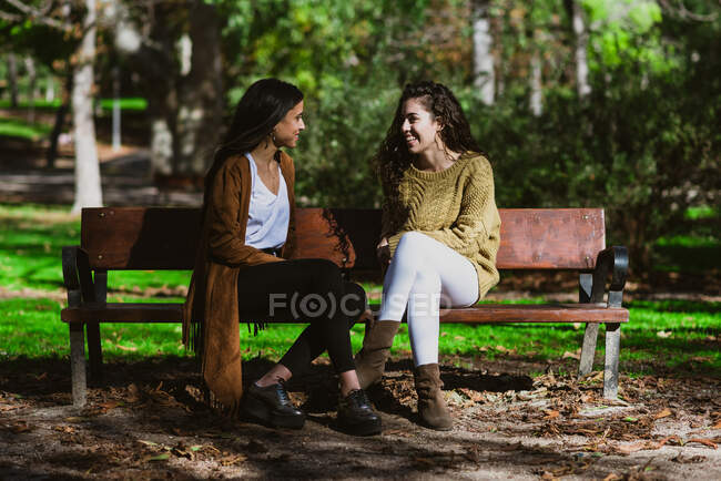 Two young beautiful female friends sitting on bench in park and chatting. — Stock Photo