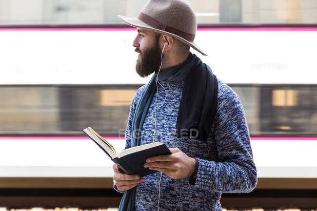 Бородатий чоловік з книгою в руках слухає музику і дивиться на залізничну станцію — стокове фото