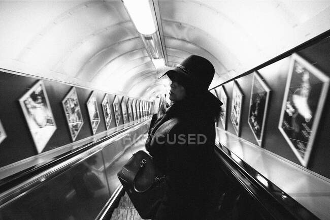 Black and white portrait of turned back woman wearing coat and hat going down on escalator in subway with pictures on walls. — Stock Photo