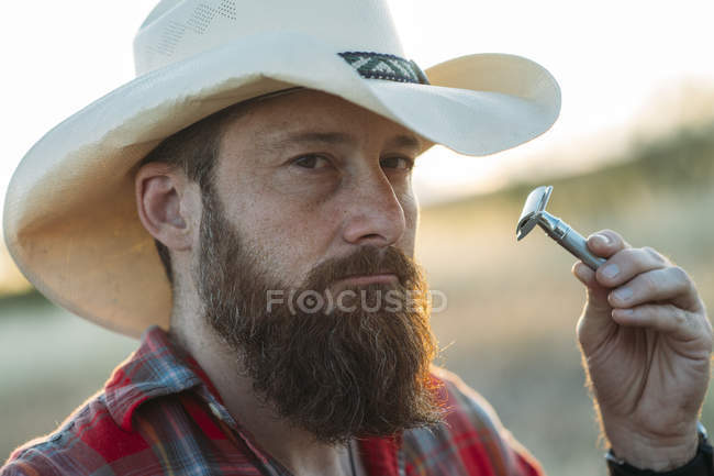 Портрет бородатого человека в ковбойской шляпе, бреющегося винтажной двойной бритвой — стоковое фото