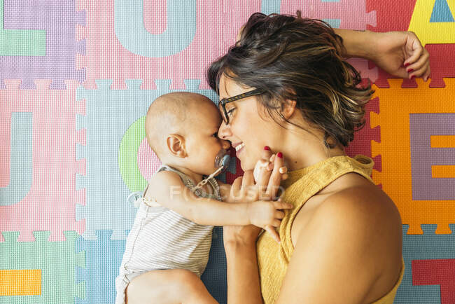 Above view of smiling brunette mother in glasses holding her baby's hand  while lying together on colorful floor of child room — Stock Photo