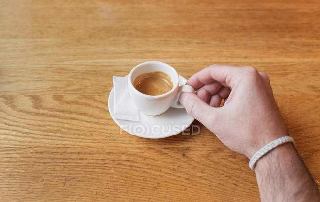 Tiro de colheita de mão masculina segurando pequena xícara branca de café expresso na mesa de madeira . — Fotografia de Stock