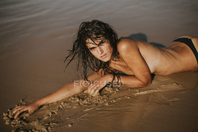 Retrato de mulher de topless com cabelo molhado deitado na areia e olhando para a câmera — Fotografia de Stock