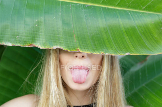Retrato de mujer rubia joven cubriendo los ojos con hoja de palmera y muestra la lengua en la cámara - foto de stock