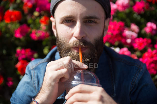 Vista frontal del hombre barbudo bebiendo batido con paja y mirando a la cámara - foto de stock