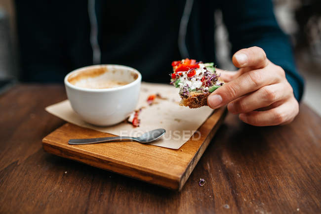 Close up view of male hand holding toast with cream and vegetables filling over wooden board with cup of cappuccino — Stock Photo