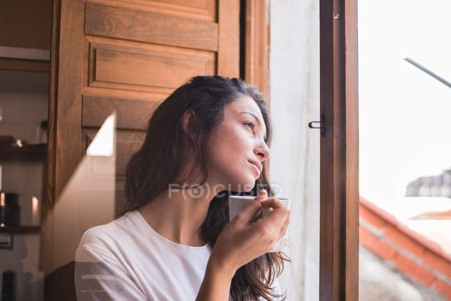 Nachdenkliche junge Frau hält Tasse in der Hand und blickt auf Fenster — Stockfoto