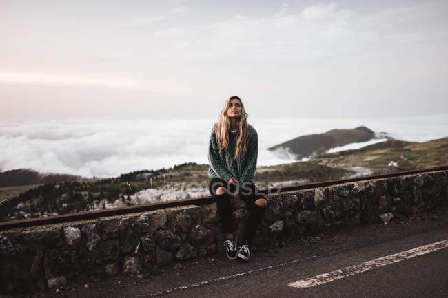 Mulher loira bonita sentada na cerca na beira da estrada em montanhas . — Fotografia de Stock