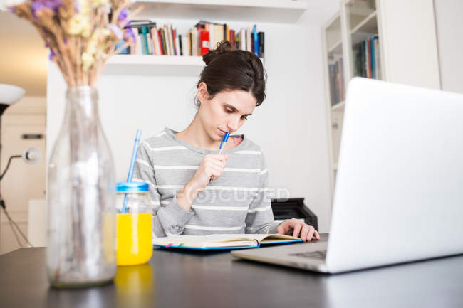 Oberflächenansicht eines nachdenklichen Mädchens, das mit Laptop am Tisch sitzt und auf das Notizbuch schaut — Stockfoto