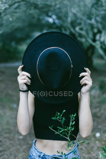 Крупный план неузнаваемой стильной девушки, стоящей на улице и держащей черную шляпу перед лицом. — стоковое фото