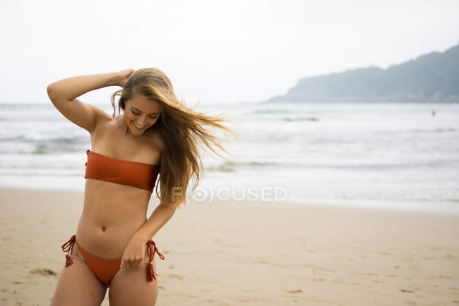 Игривая девушка в бикини позирует на песчаном пляже и смотрит вниз — стоковое фото