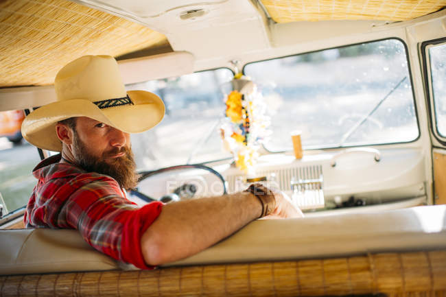 Bearded man in cowboy hat sitting at drivers seat of retro van and looking over shoulder at camera — Stock Photo