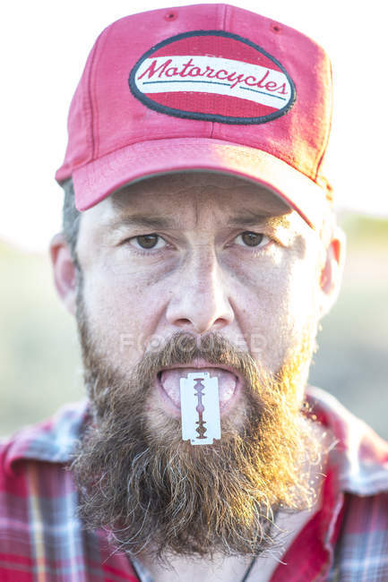 Portrait of bearded man in cap holding razor on tongue and looking at camera — Stock Photo