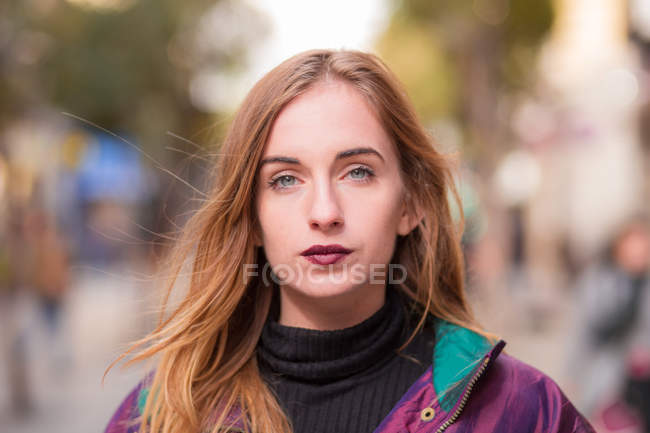 Jeune fille élégante avec rouge à lèvres sombre regardant la caméra sur fond de rue floue . — Photo de stock