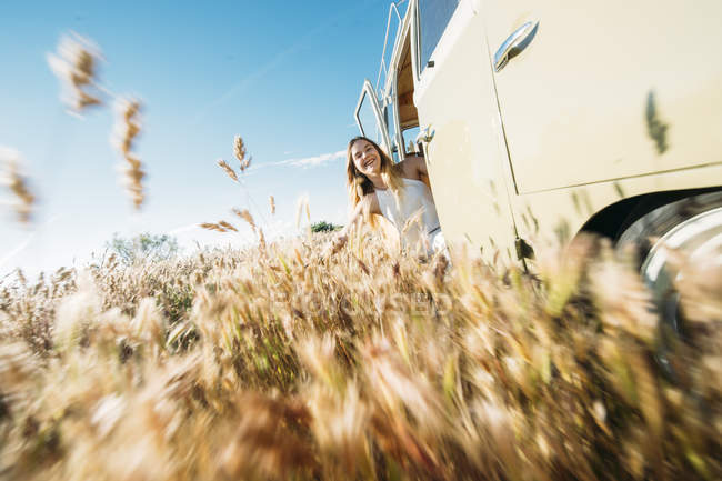 Menina alegre inclinando-se para fora da porta da van e tocando centeio no campo — Fotografia de Stock