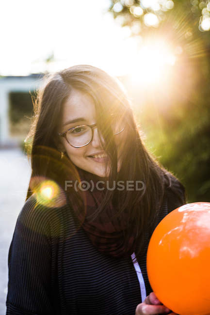Junge Brünette mit Brille hält Luftballon und lächelt im Sonnenlicht in die Kamera. — Stockfoto