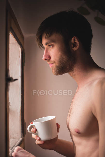 Giovane uomo assonnato senza camicia che tiene la tazza e guarda la finestra — Foto stock