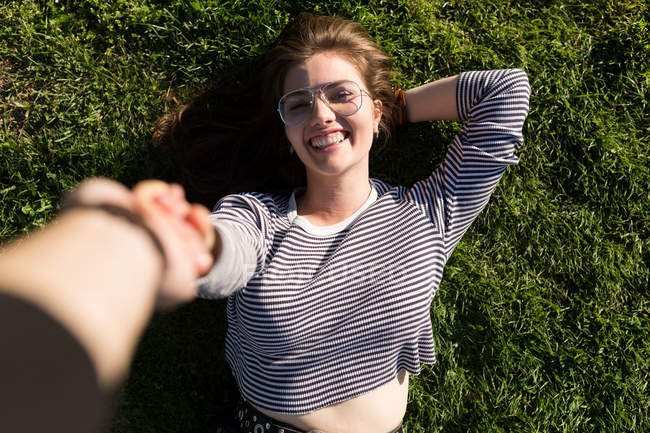 From above shot of cheerful  girl in glasses lying on grass holding hand of photographer. — Stock Photo