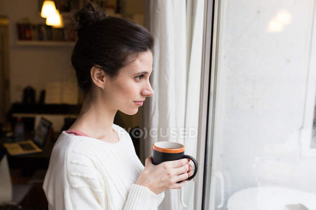 Vista lateral de mujer morena posando con taza de cacao y mirando a la ventana - foto de stock
