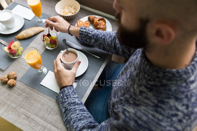 Зверху бородатий чоловік їсть фруктовий салат і тримає чашку кави за журнальним столиком — стокове фото