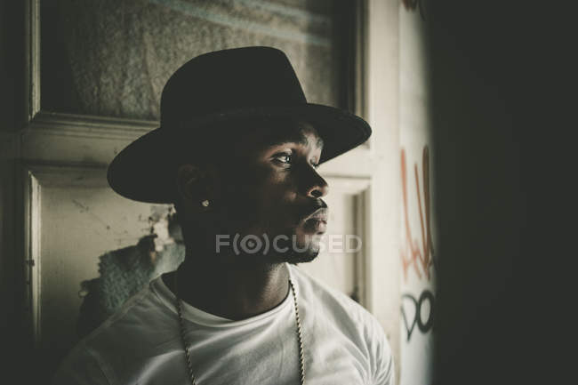 Hombre africano en sombrero en habitación oscura abandonada con graffiti en la pared. Mirando hacia otro lado . - foto de stock
