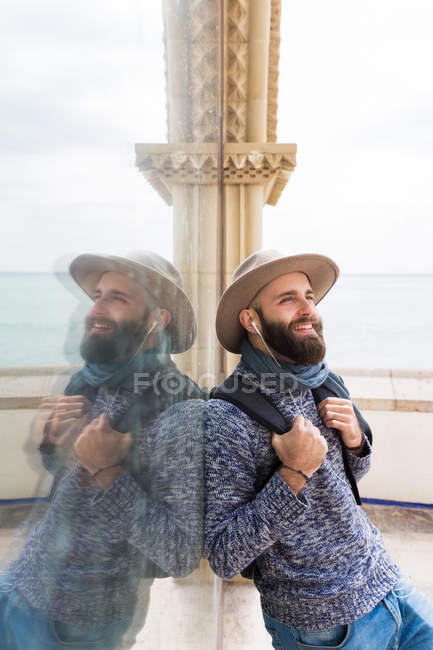 Retrato de hombre barbudo sonriente con sombrero apoyado en vidrio y escuchando música
 . - foto de stock