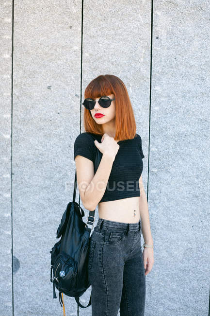 Руда дівчина з червоними губами позує в сонцезахисних окулярах над сірою стіною — стокове фото