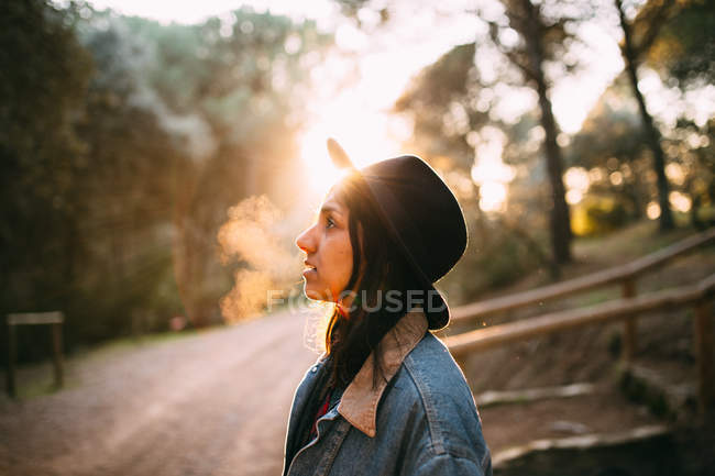 Vista lateral da jovem mulher em chapéu na estrada iluminada pelo sol na floresta — Fotografia de Stock