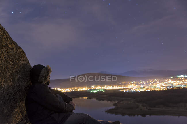Hombre en la cima de una montaña y disfrutando de la vista del cielo nocturno y las luces de la ciudad - foto de stock